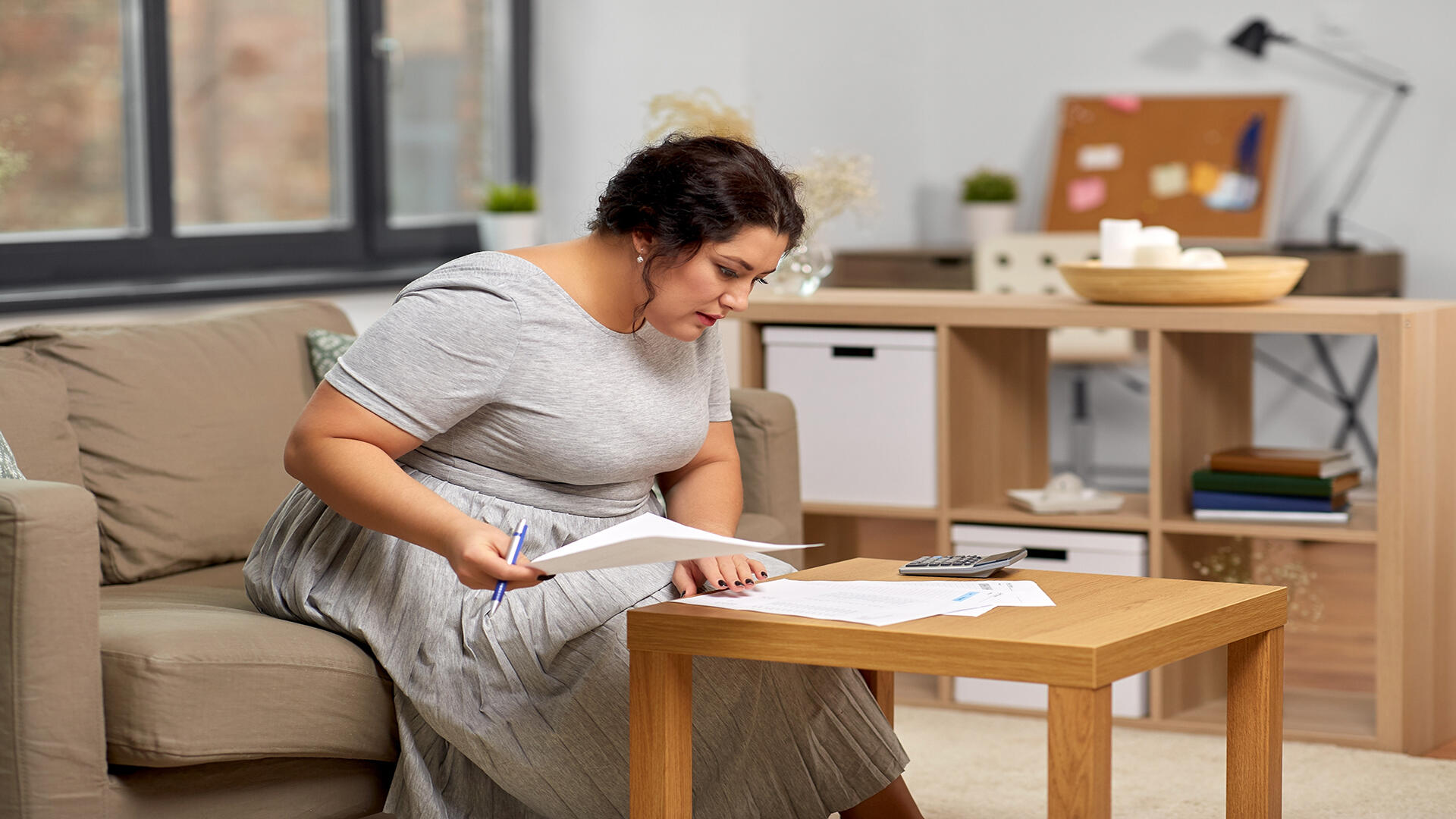 A focused plus-size businesswoman calculating finances with a pen and paper at her cozy home office, featuring a wooden coffee table, a calculator, and an organized bookshelf in the background.