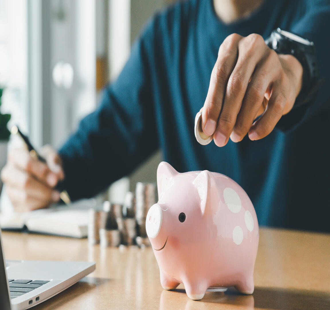 Person in a navy blue sweater saving money by inserting a coin into a pink polka-dotted piggy bank, with a laptop and notepad in the foreground, symbolizing financial planning and savings.